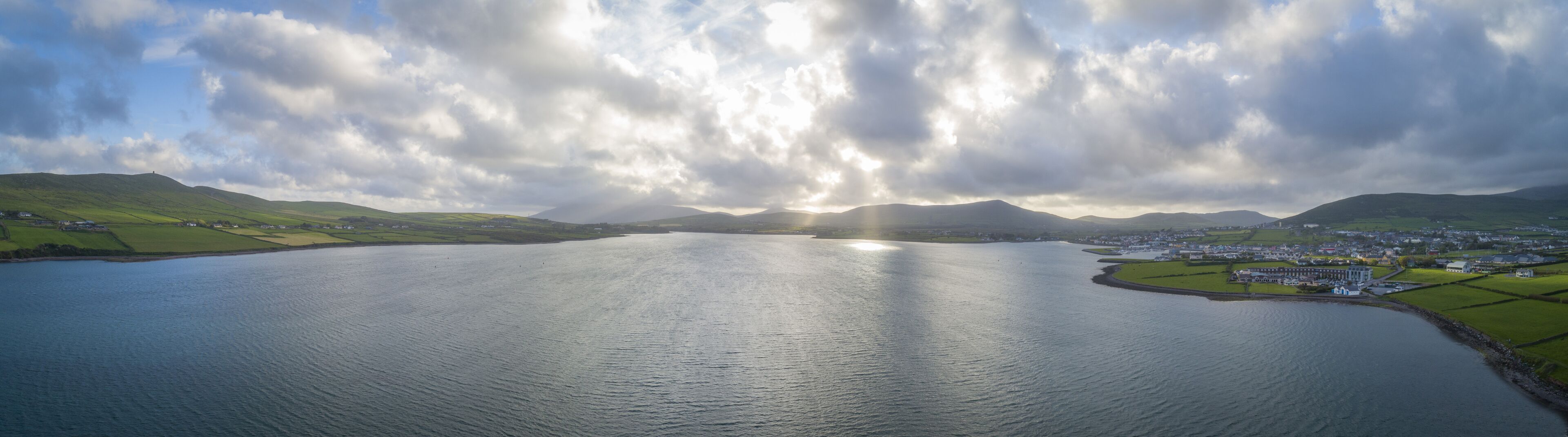 A coastal town with a large body of water, green hills, and a partly cloudy sky.