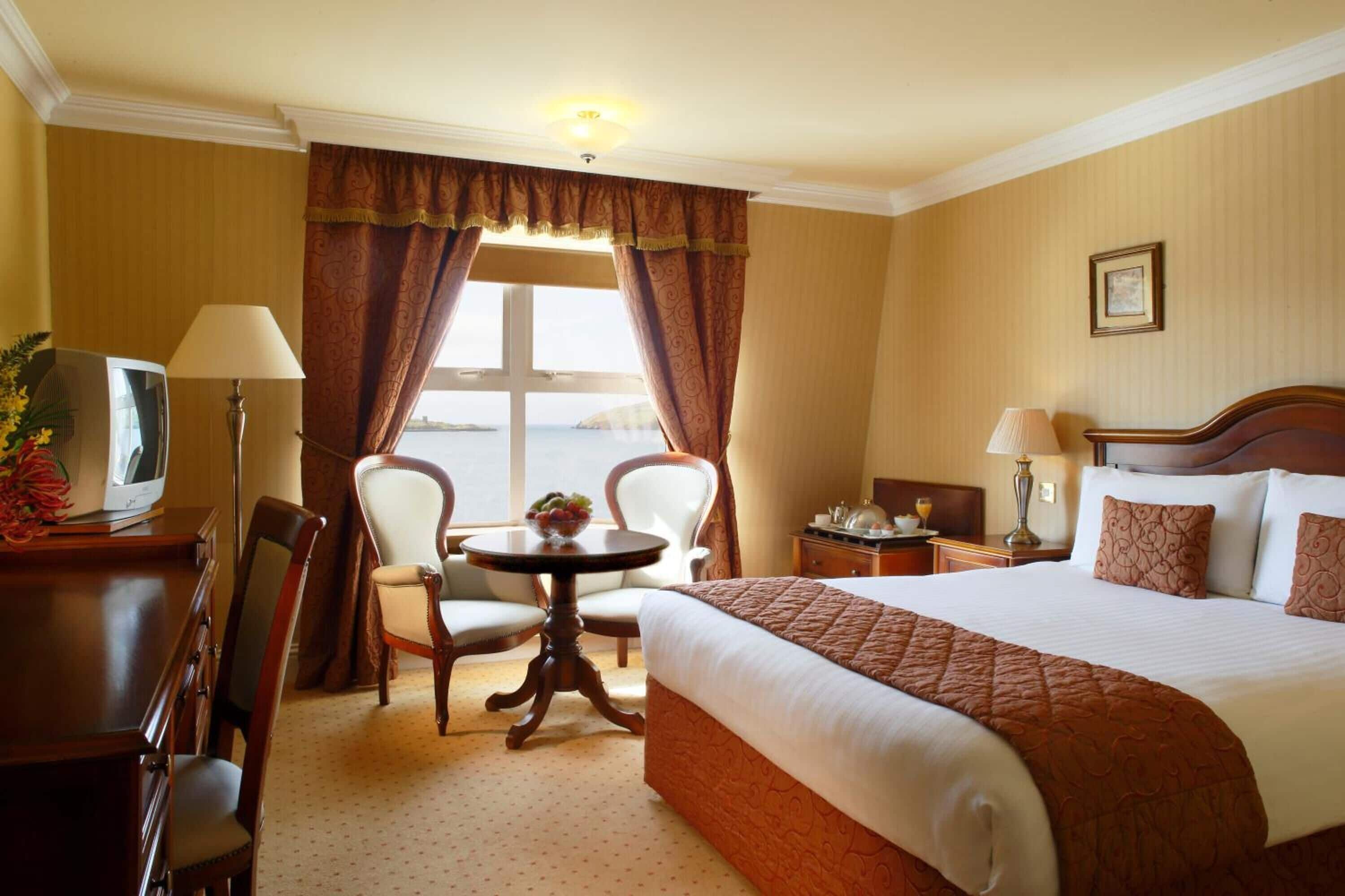 Ein Hotelzimmer mit einem großen Bett, zwei Stühlen, einem kleinen Tisch, einem Fernseher und einem Fenster mit Vorhängen.