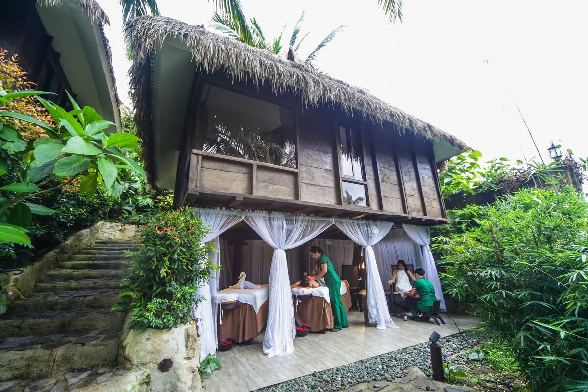 A traditional wooden building with thatched roofs, a stone staircase, and a small outdoor seating area with a table and chairs.