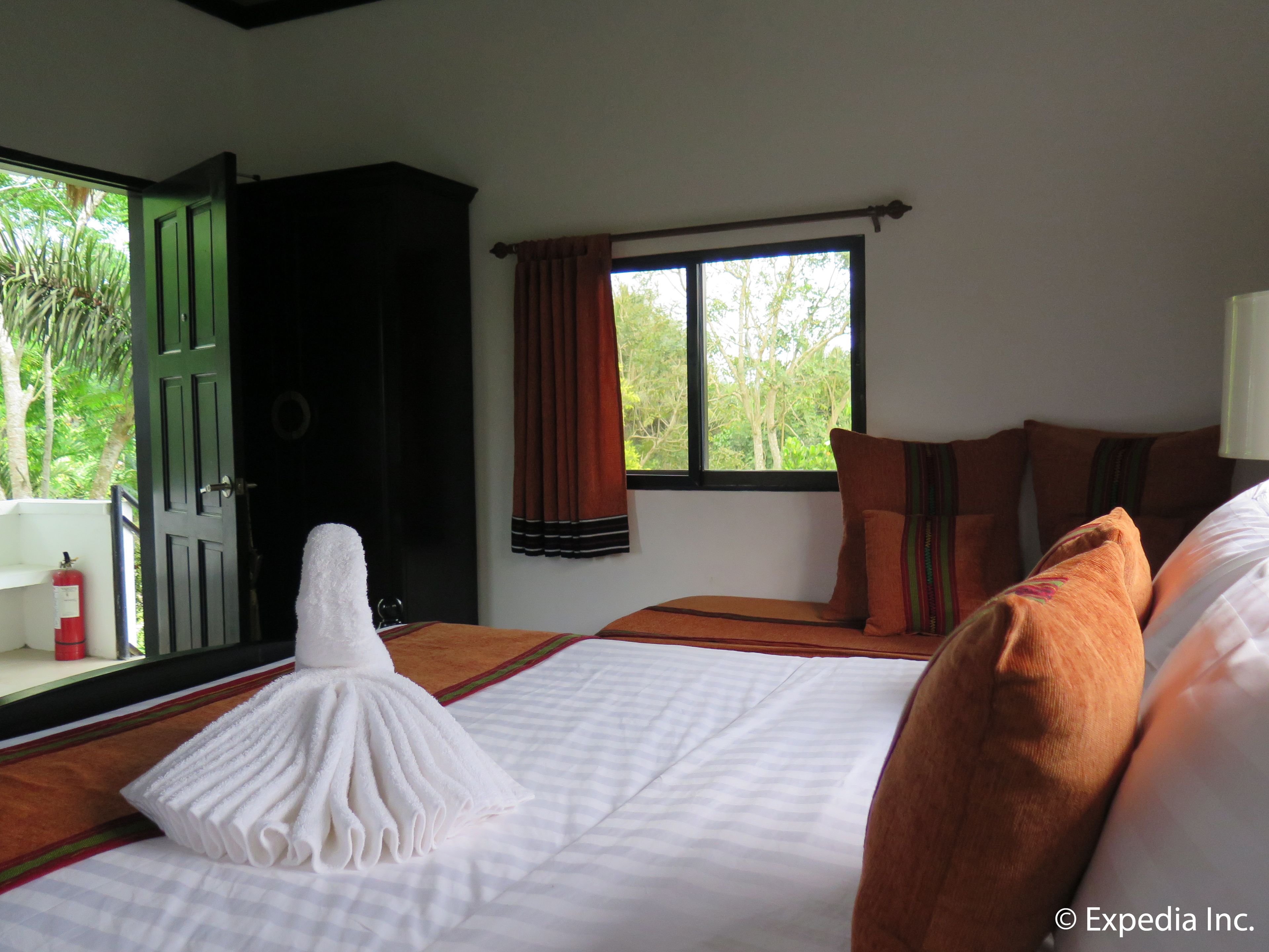 A hotel room with a bed, a window with curtains, a door, and a view of greenery outside.