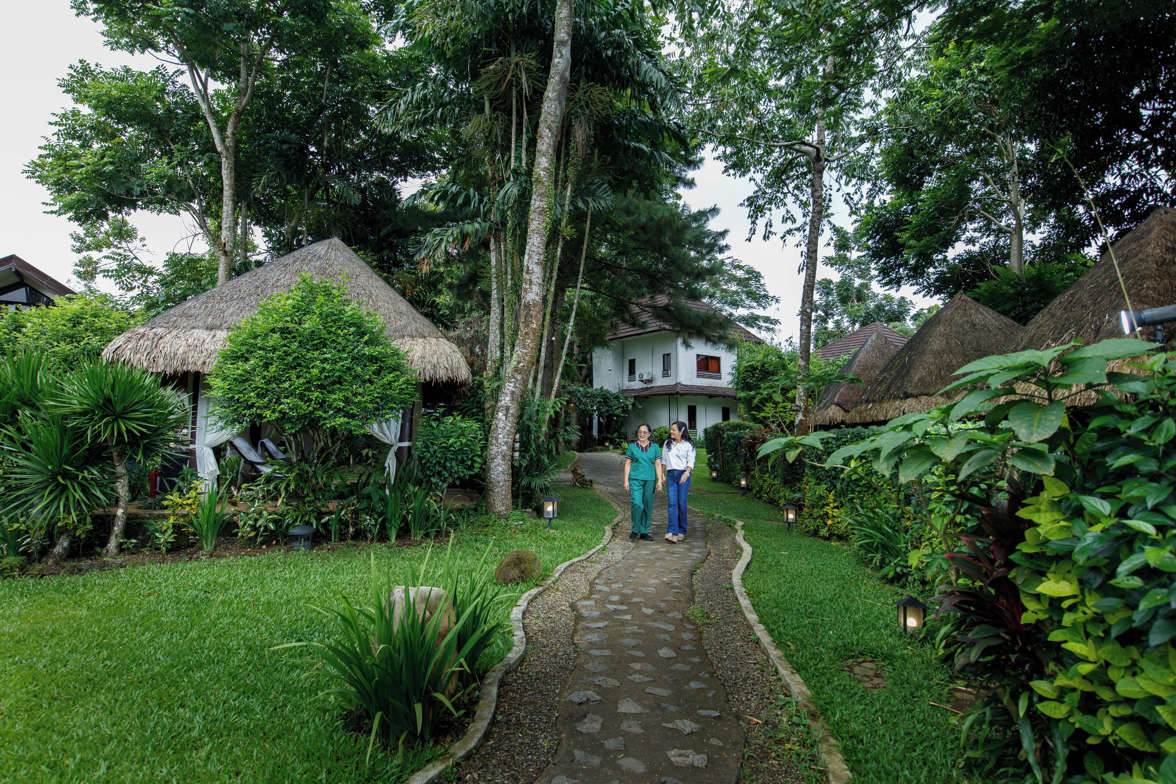 A pathway leading to a house surrounded by lush greenery and thatched-roof structures.