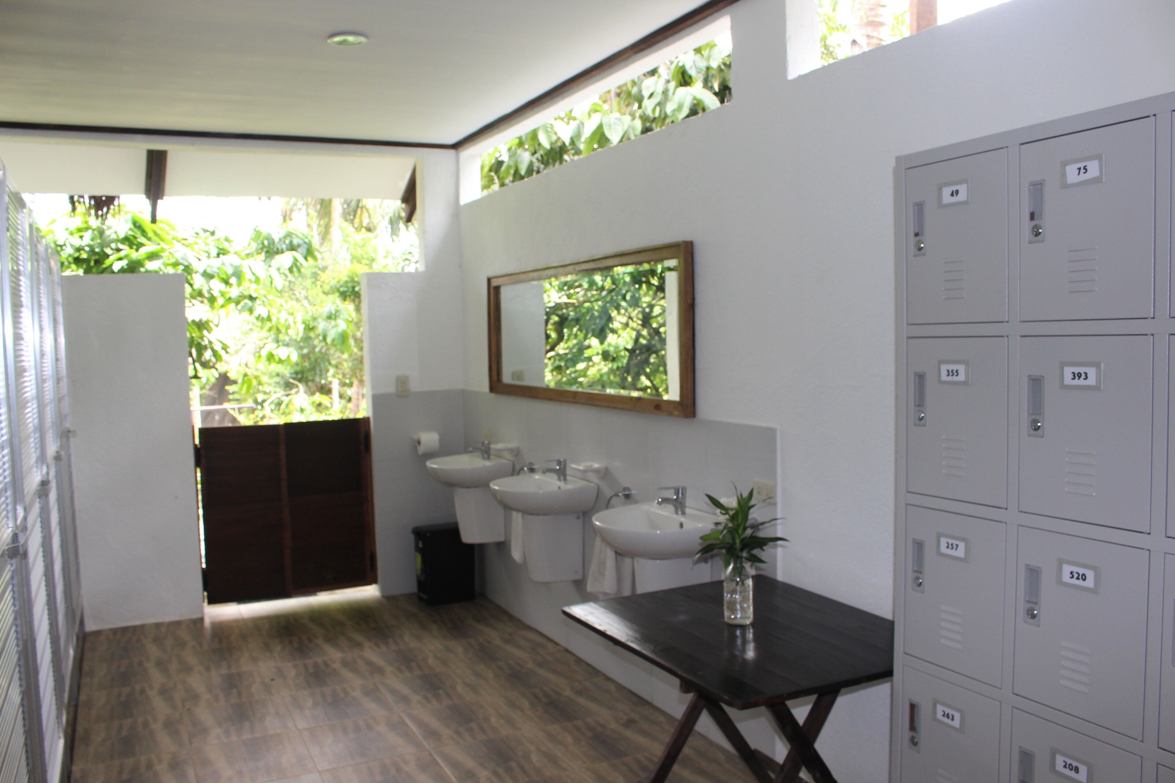 A bathroom with two sinks, a mirror, and a potted plant. There are lockers and a trash bin.