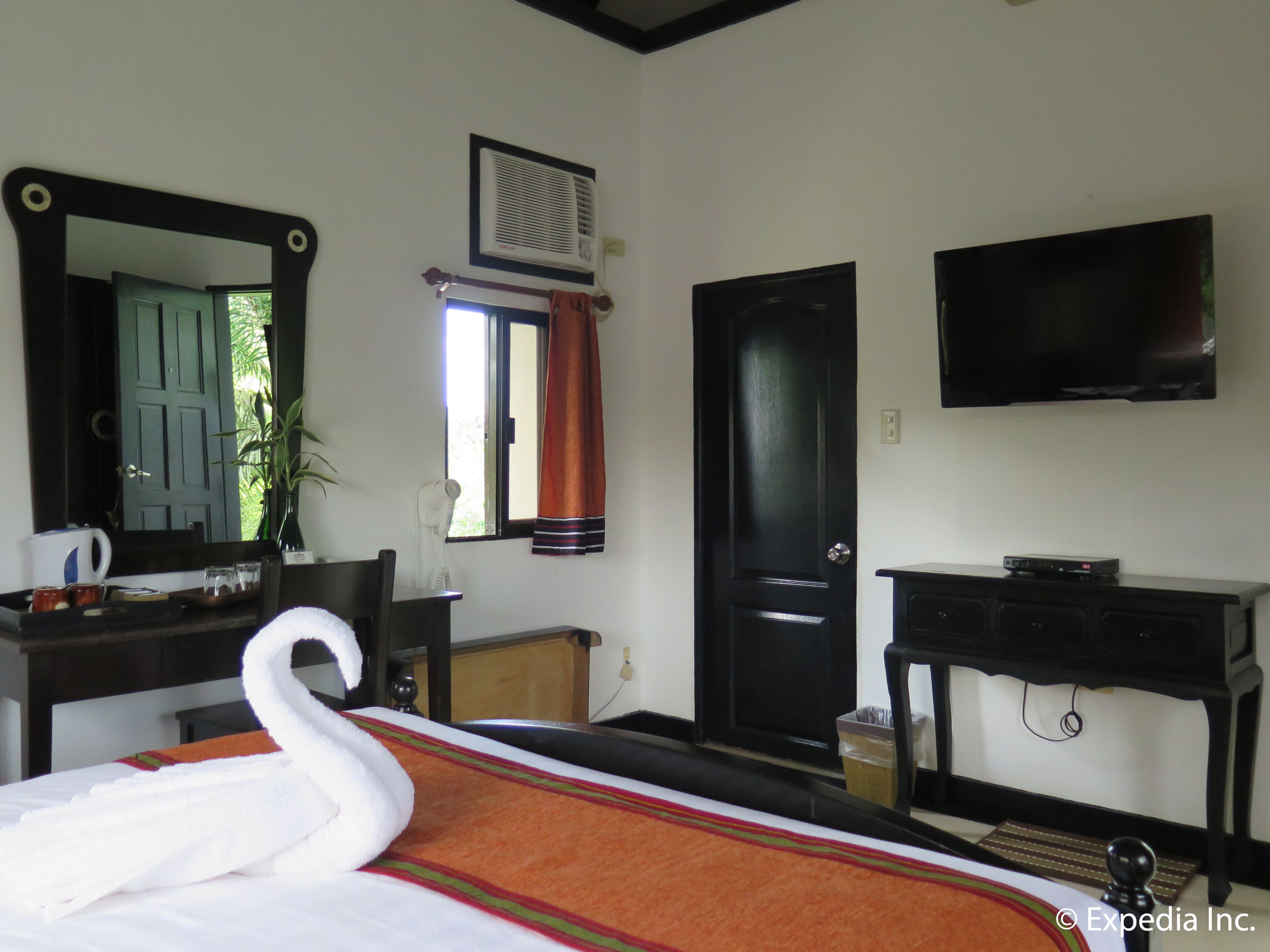 A hotel room with a bed, a TV, a desk, a chair, a mirror, a window with curtains, and a plant.