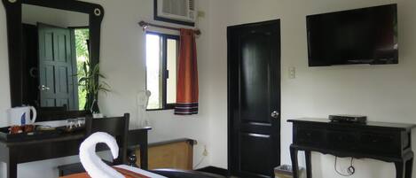 A hotel room with a bed, a TV, a desk, a chair, a mirror, a window with curtains, and a plant.