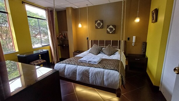A hotel room with a bed, bedside table, and a desk with a chair. There is a window with curtains, a hanging light fixture, and a wall-mounted telephone. - Wifala Thematic Hotel (Cusco)