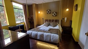 Habitación de hotel con cama, mesita de noche y escritorio con silla. Se ve una ventana con cortinas, una lámpara colgante y un teléfono instalado en la pared.