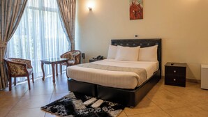 A hotel room with a bed, a chair, a small table, a nightstand, and a painting on the wall. - Yaya Africa Athletics Village (Sululta)