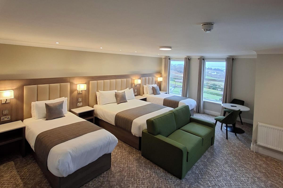 Une chambre d’hôtel avec deux lits, un canapé vert, une petite table et une vue sur l’extérieur.