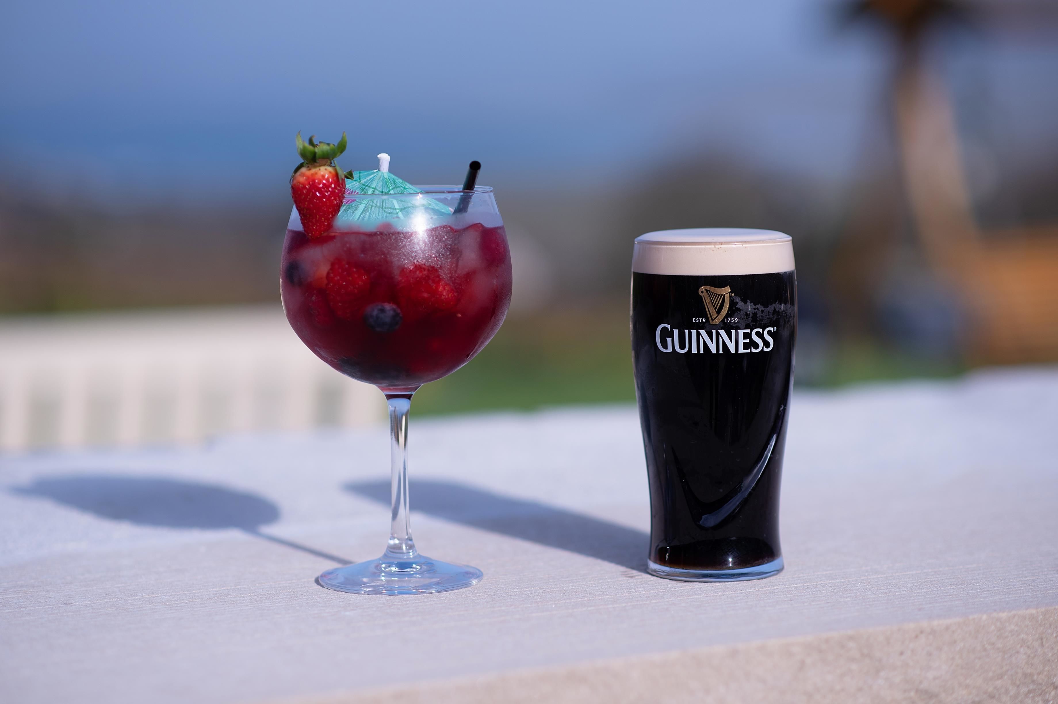 Un vaso de bebida roja con una fresa y un paraguas azul sobre una mesa. Junto a él, un vaso de Guinness.