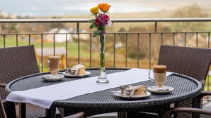 A round wicker table with a white tablecloth, two cups of coffee, a vase with flowers, and plates with desserts on a balcony overlooking a landscape.