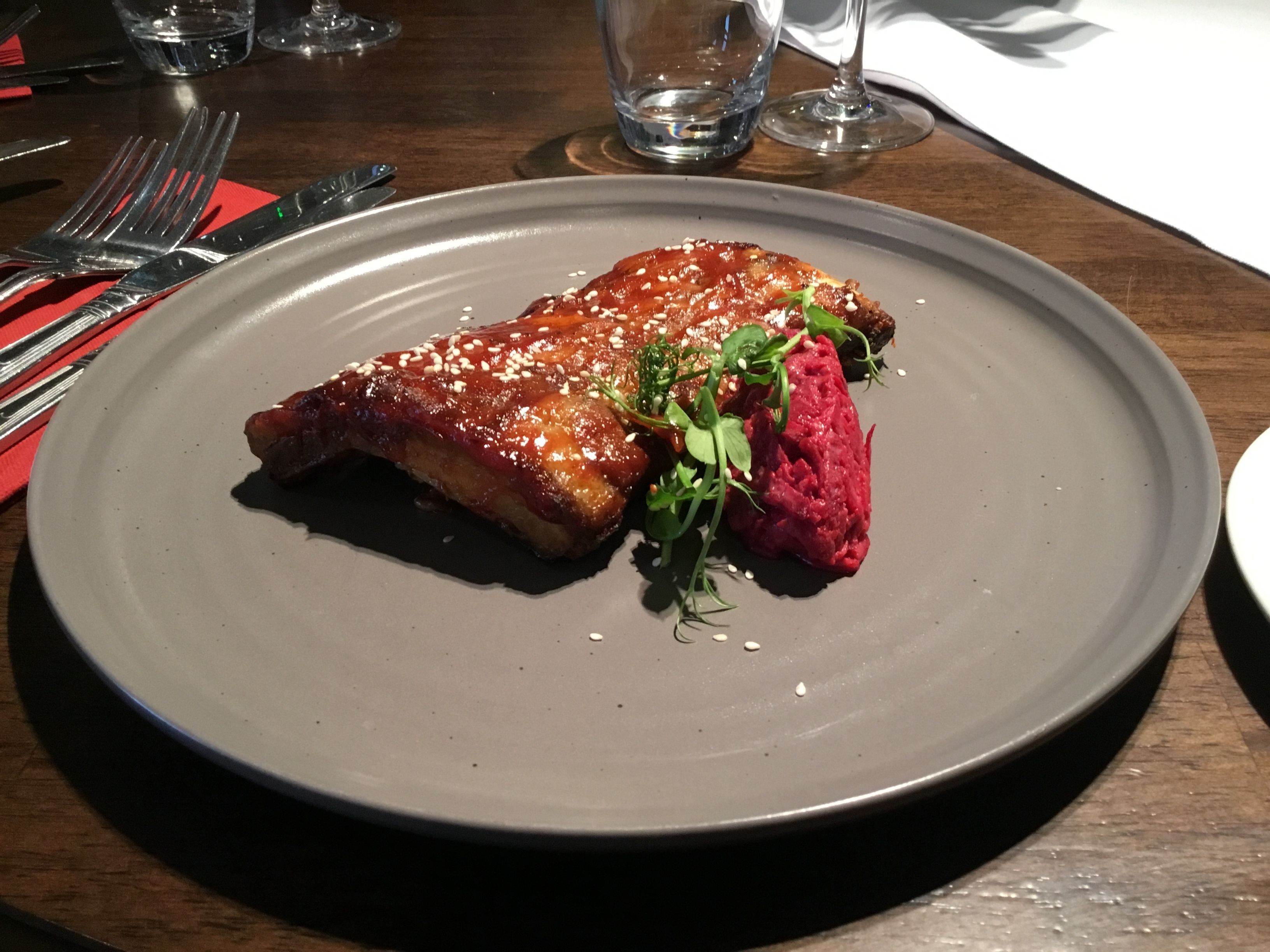 Ein Teller mit einem glasierten Fleischgericht und einer Beilage aus roter Sauce, platziert auf einem dunklen Tisch, im Hintergrund sind Besteck und Gläser zu sehen.