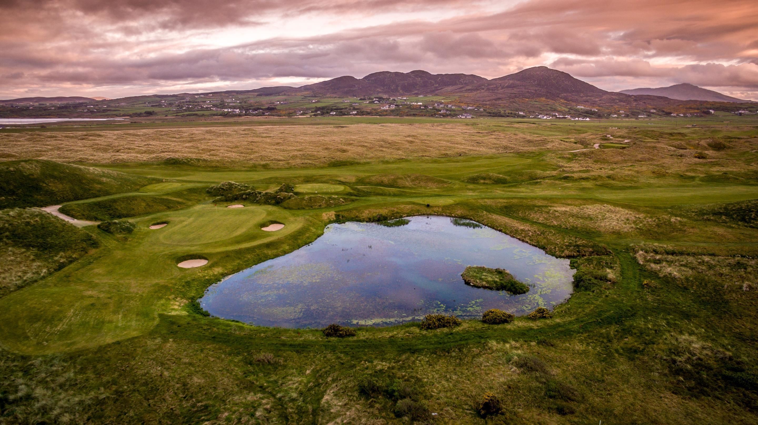 Un parcours de golf avec un étang, entouré de prairies verdoyantes et de montagnes en arrière-plan.