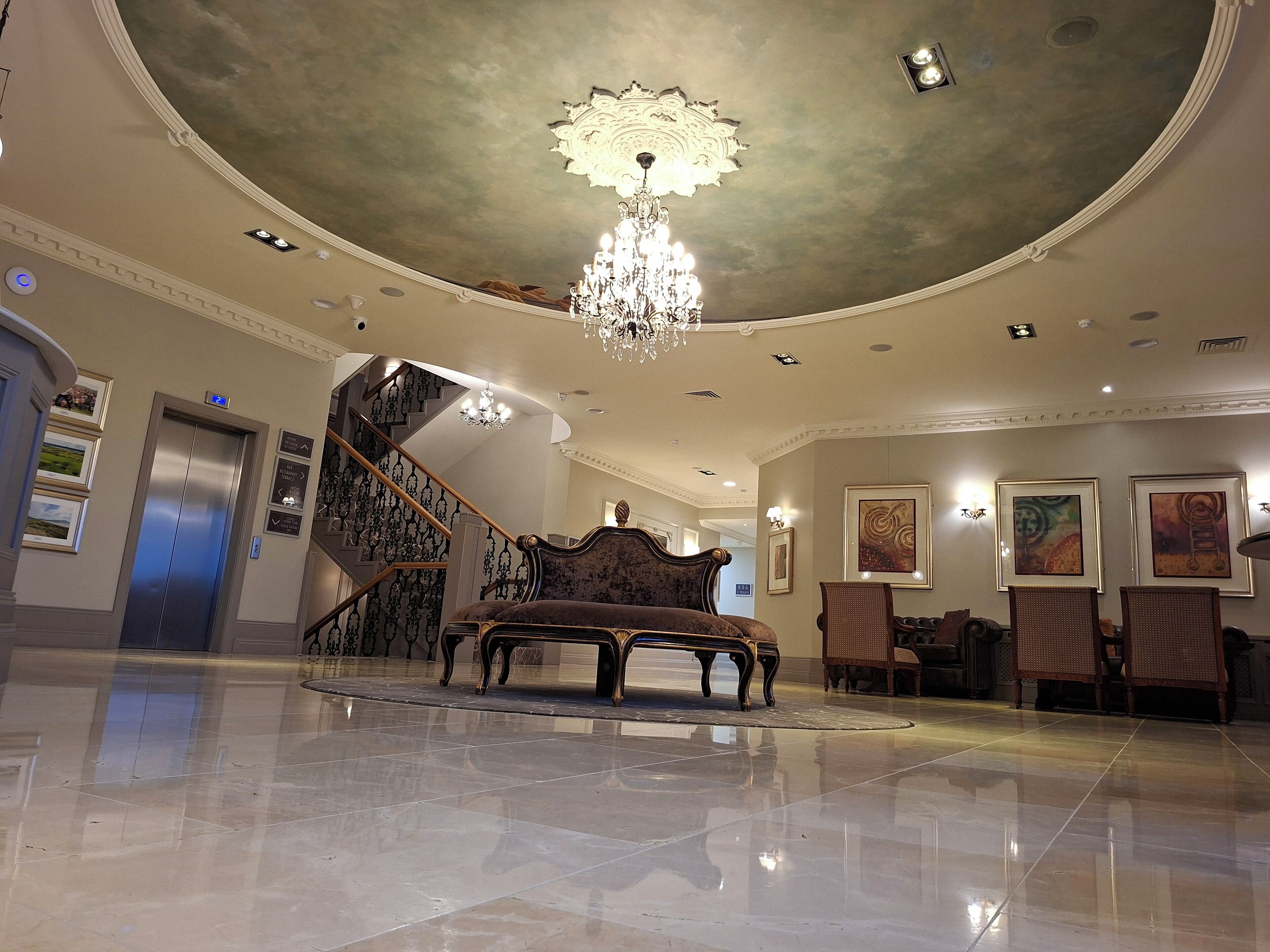 A spacious lobby with a grand chandelier, a plush bench, and a staircase leading to an upper floor.
