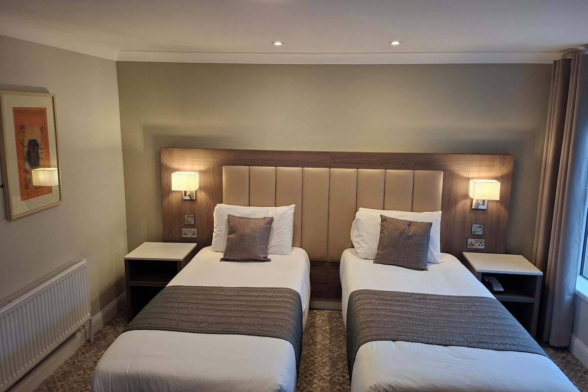 Une chambre d’hôtel avec deux lits, une tête de lit, des lampes de chevet, un tableau encadré et un radiateur.