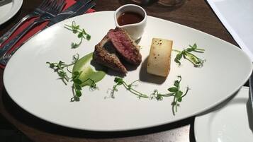 Un plato blanco con un trozo de carne, un cubito de queso y una taza pequeña de salsa, adornado con hierbas verdes.