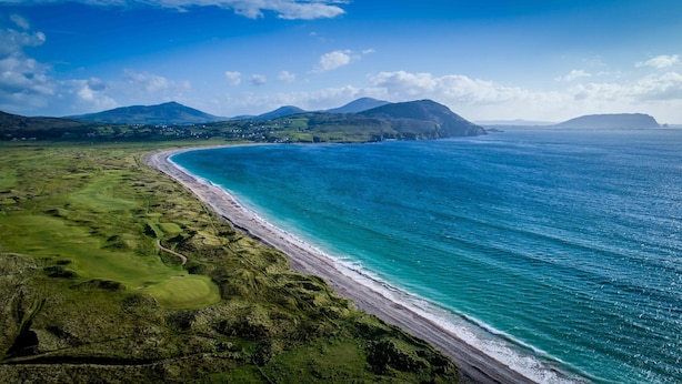 A coastal landscape with a beach, green fields, and distant mountains.