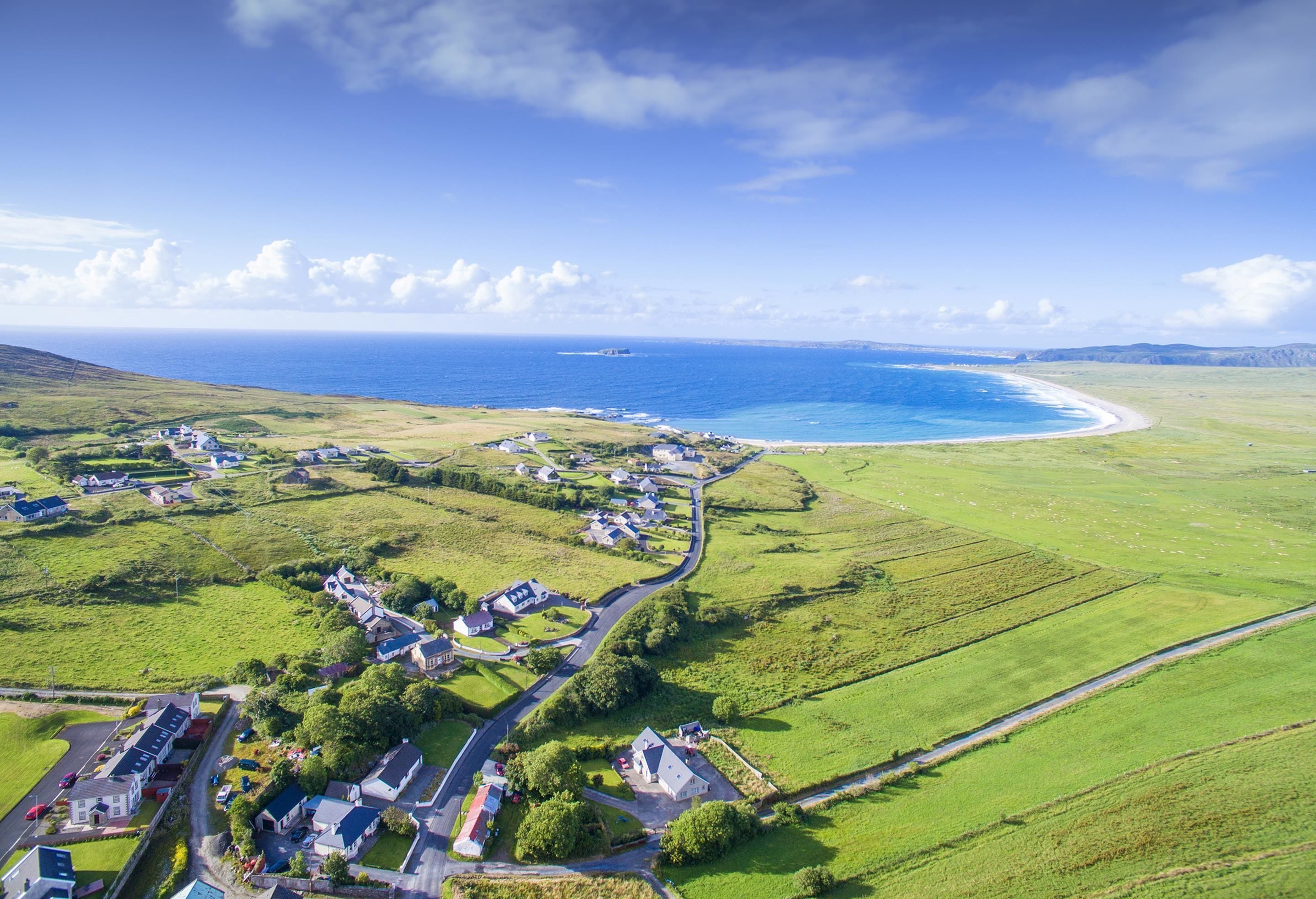 A coastal village with houses along a curved road, a beach, and a clear blue sky.