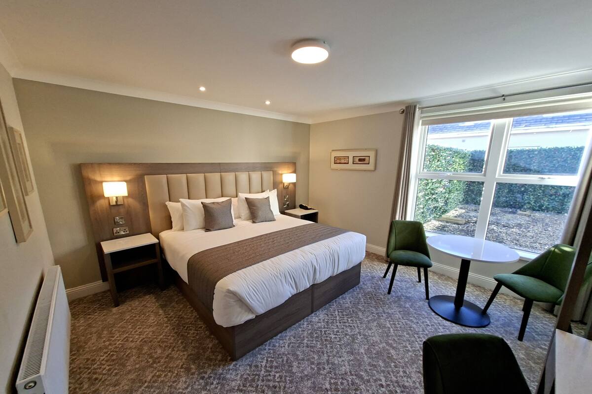 Une chambre d’hôtel avec un grand lit, deux tables de chevet, un bureau avec une chaise et une fenêtre donnant sur un paysage verdoyant.