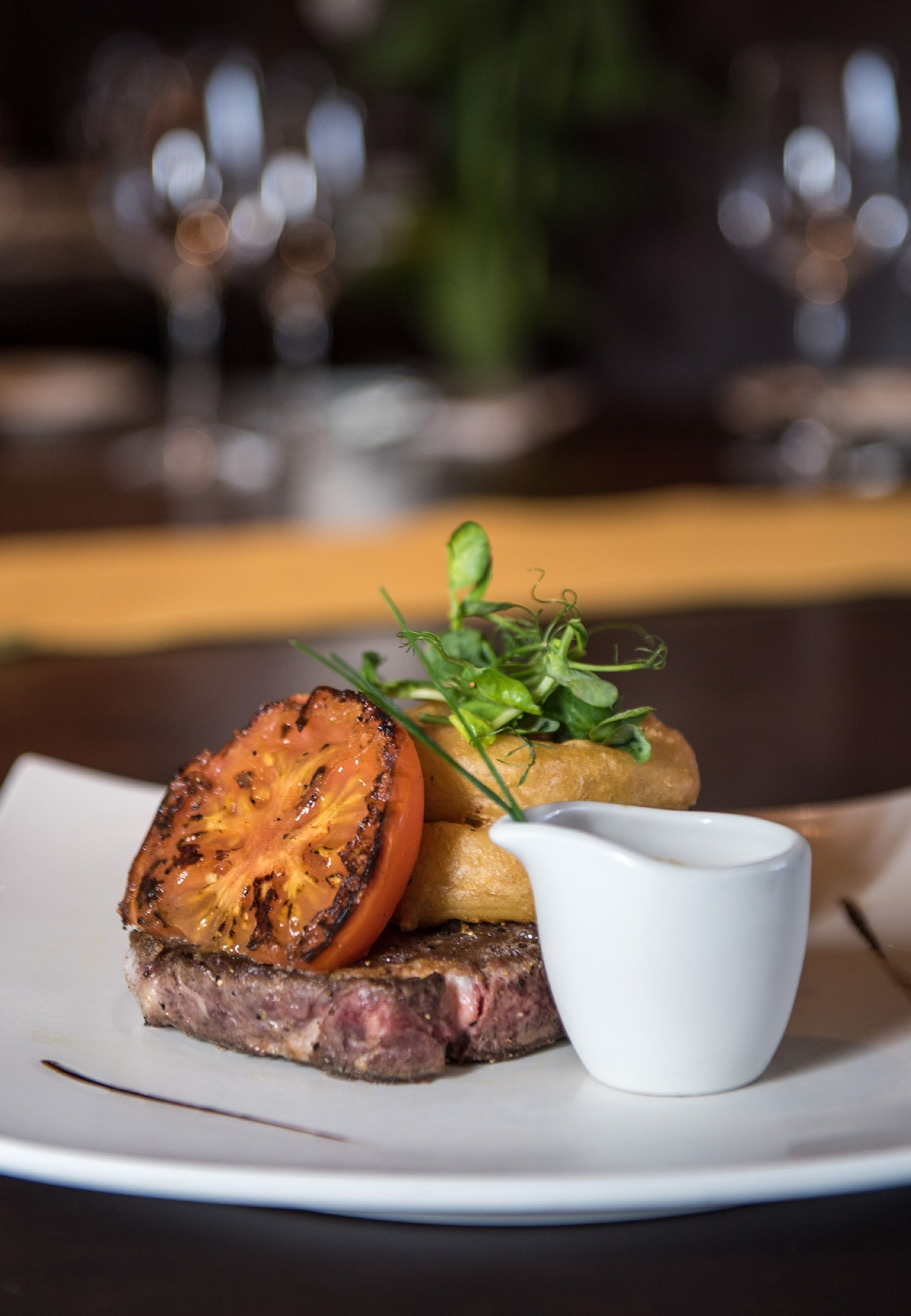 A steak with a slice of tomato and greens on a plate, accompanied by a small white cup, likely containing sauce.