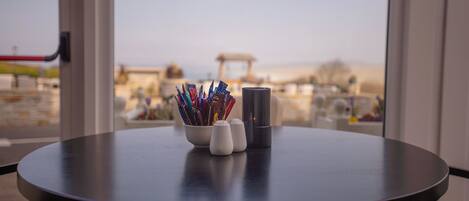 Una mesa redonda con un tazón de bolígrafos, saleros y pimenteros, un vaso negro, y vistas a una playa y edificios a través de amplios ventanales.