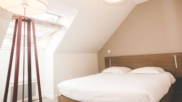 Camera da letto con soffitto a falde, finestra a lucernario, letto con biancheria bianca e lampada da terra.
