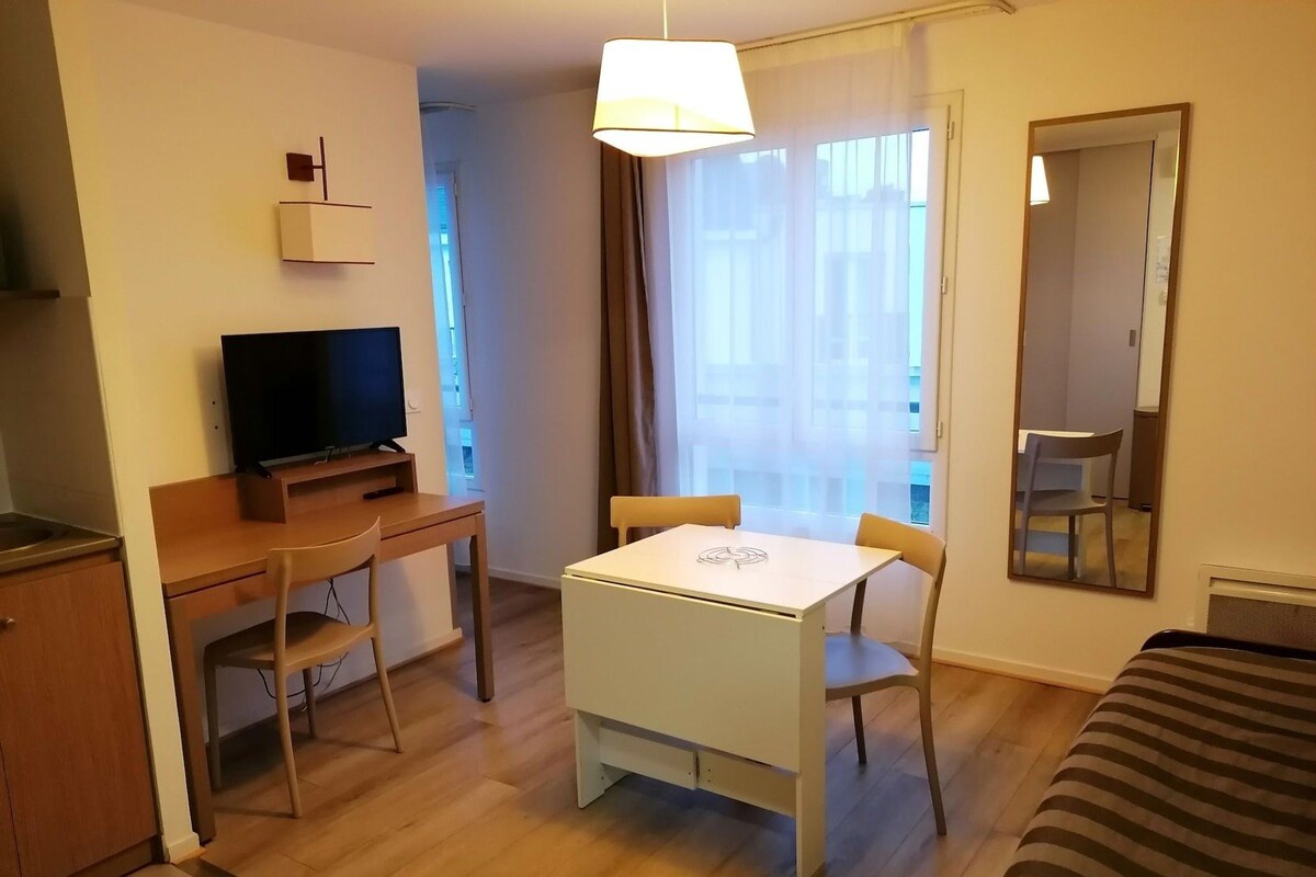 A hotel room with a bed, a desk with a TV, a dining table with chairs, a mirror, and a window with curtains.
