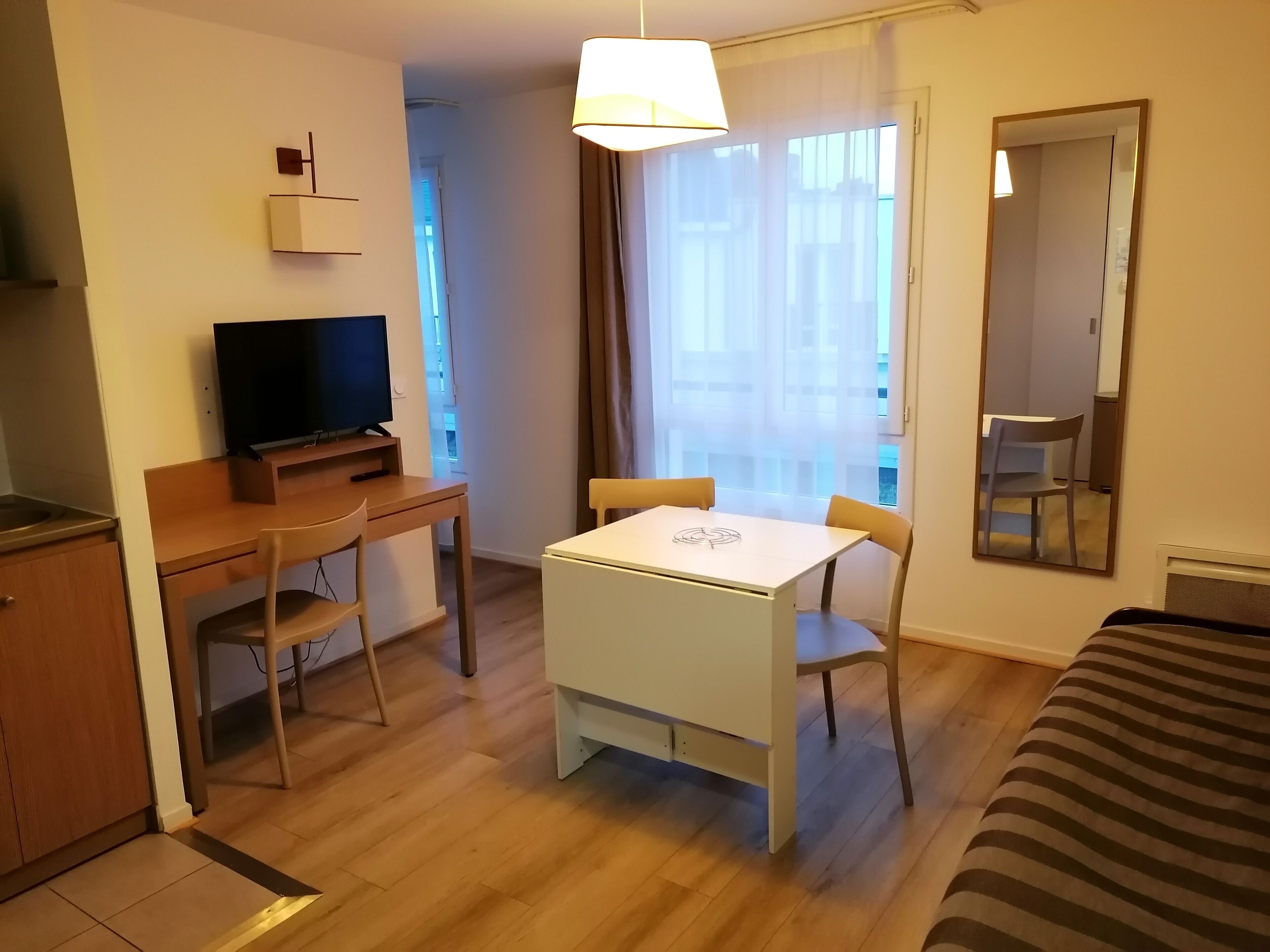 Una habitación de hotel con escritorio de madera, televisor, mesa de comedor con sillas, cama, espejo y ventana con cortinas.