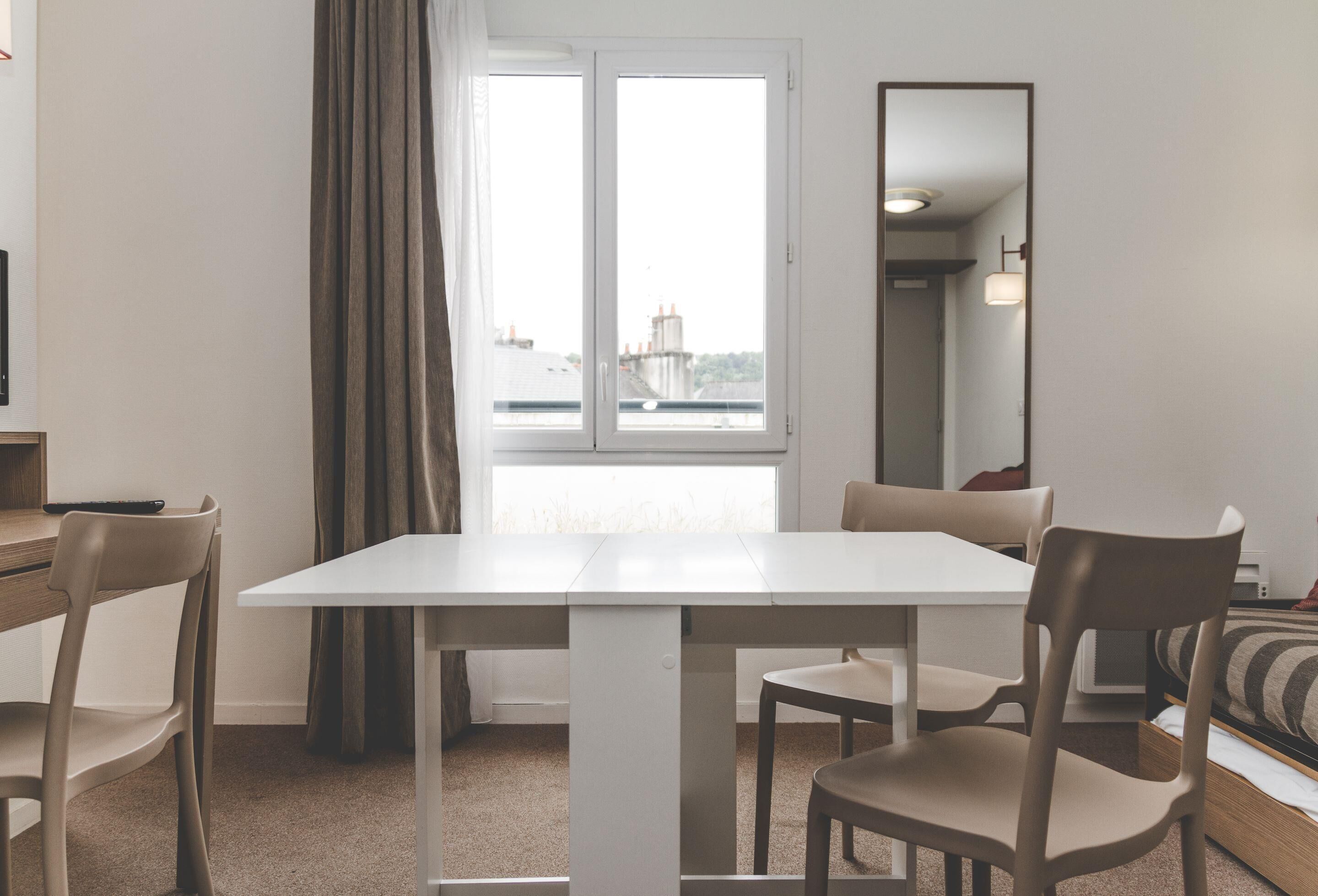 Una habitación moderna y minimalista con una mesa blanca y cuatro sillas, un ventanal con cortinas y una cama con ropa de cama a rayas.