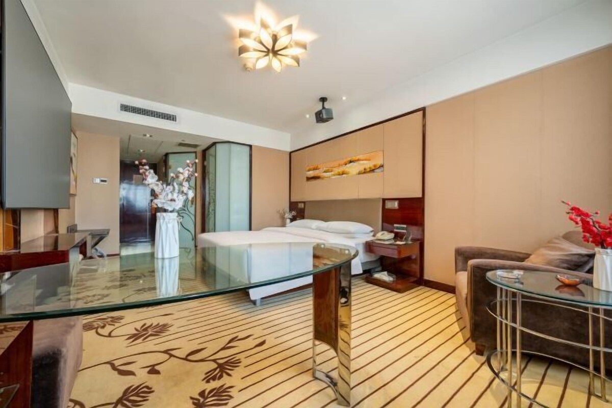 A modern hotel room with a glass dining table, a bed with white linens, a sofa, and a large window.