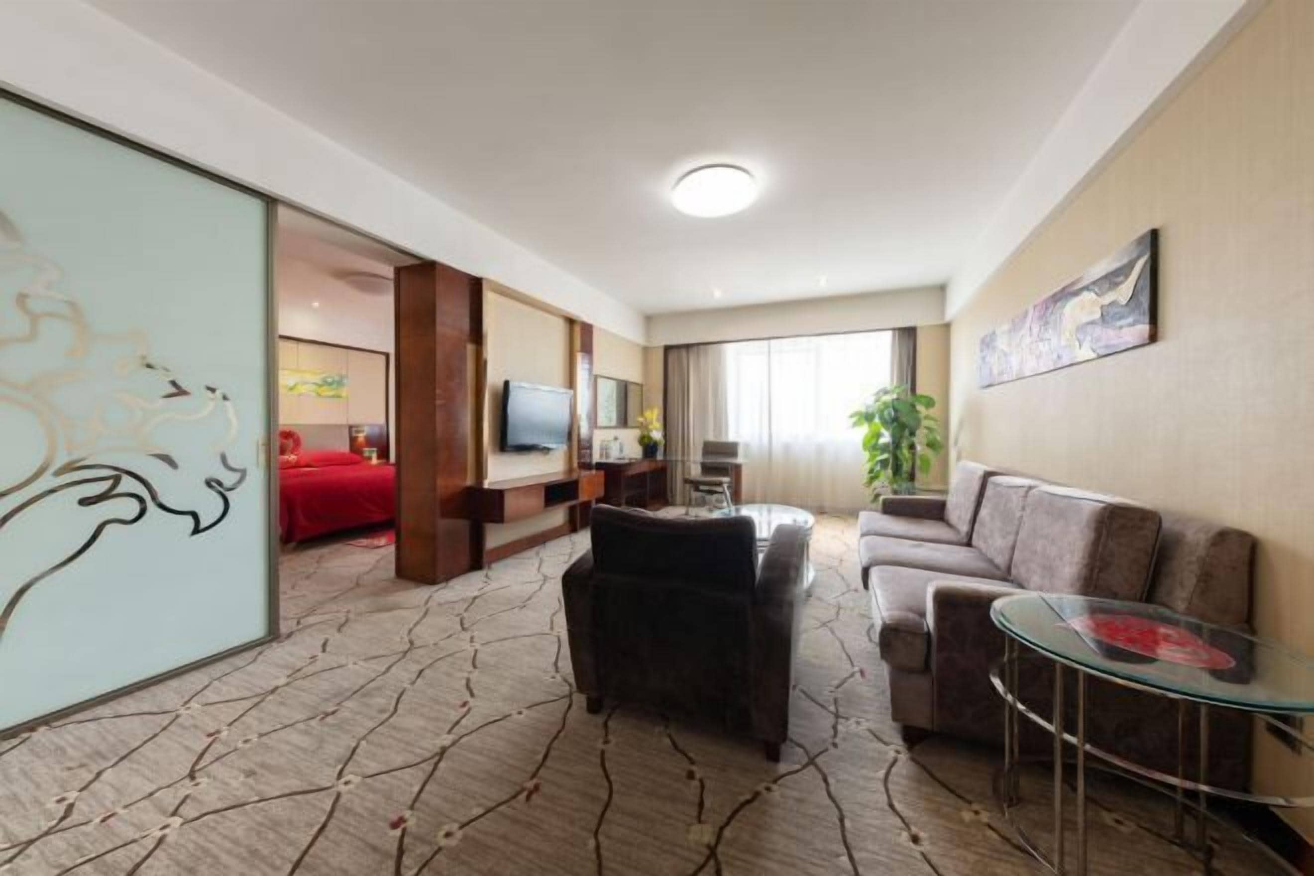 A hotel room with a living area, a sofa, a glass coffee table, a TV, and a red bed.