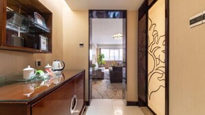 Eine Hotellobby mit einer Rezeption, einer Kaffeestation und Blick in einen Wohnbereich mit Sofa und Pflanze.