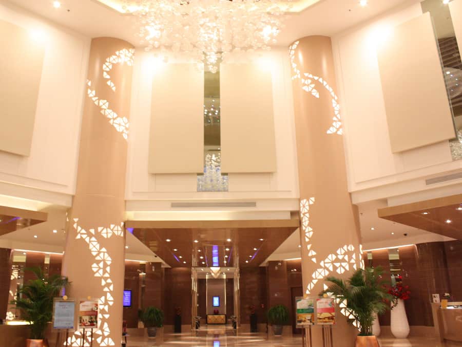 A spacious lobby with high ceilings, large columns, and a central chandelier. There are reception desks and potted plants.