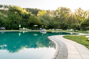 A swimming pool with a curved edge, surrounded by lounge chairs and umbrellas, with trees in the background. - Aparthotel Quinta do Crestelo (Seia)