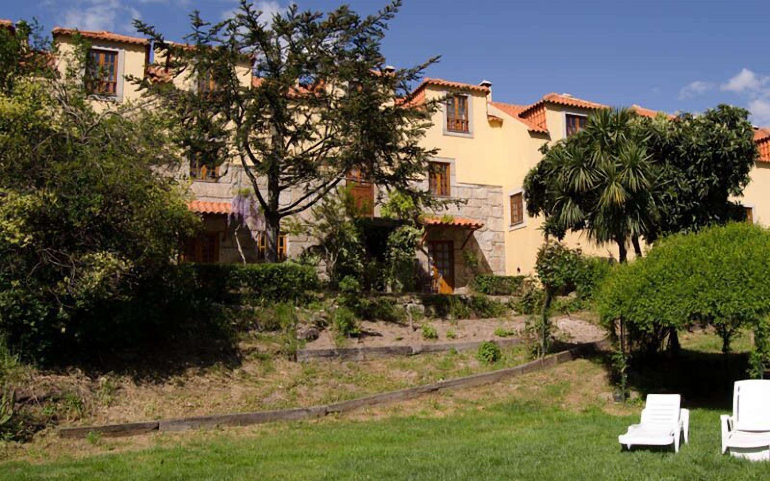 A building with multiple windows and a stone facade, surrounded by greenery and a grassy area with white chairs.