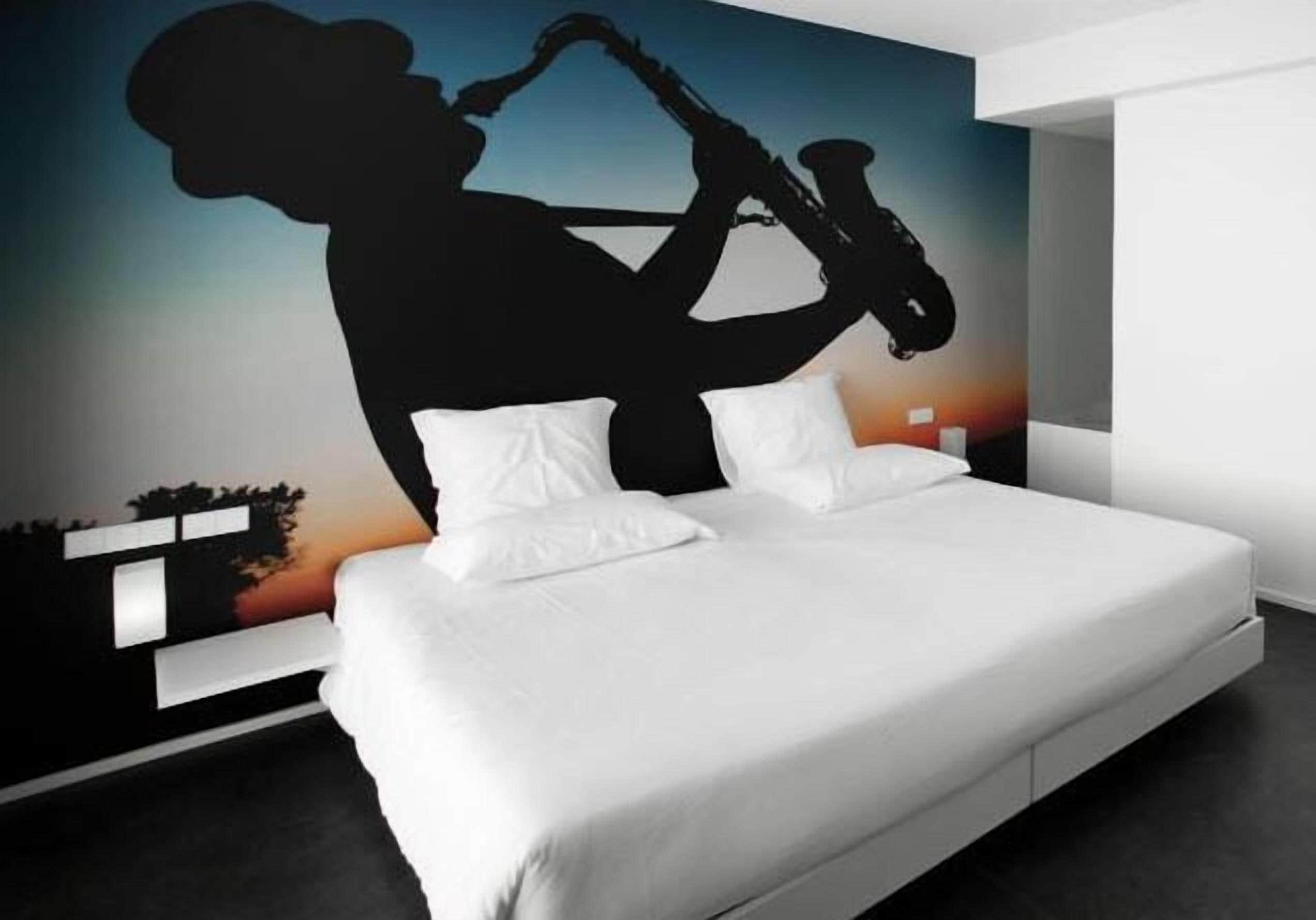 A modern bedroom with a large bed, white bedding, and a wall mural of a saxophonist.