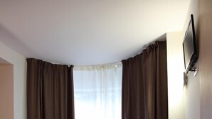 Un dormitorio con piso de madera, una cama roja, cortinas marrones y un televisor instalado en la pared.