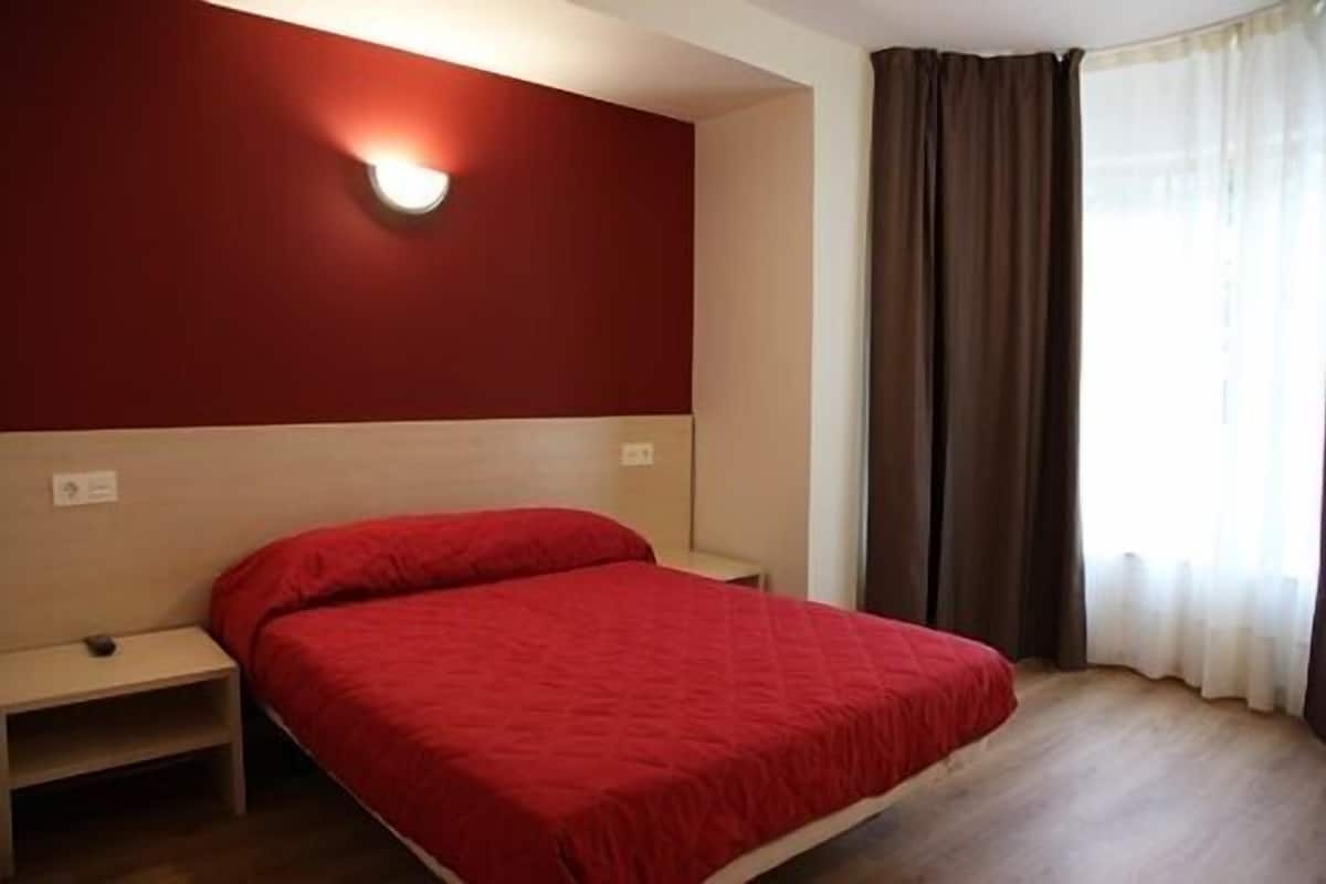 Une chambre d’hôtel avec un lit, une table de chevet, une lampe fixée au mur et une fenêtre avec des rideaux.