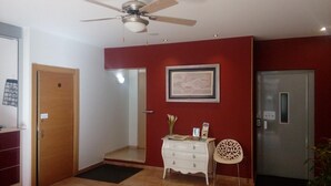 Una habitación de hotel con ventilador de techo, un cuadro enmarcado en la pared, un aparador blanco, una silla estampada y un refrigerador.