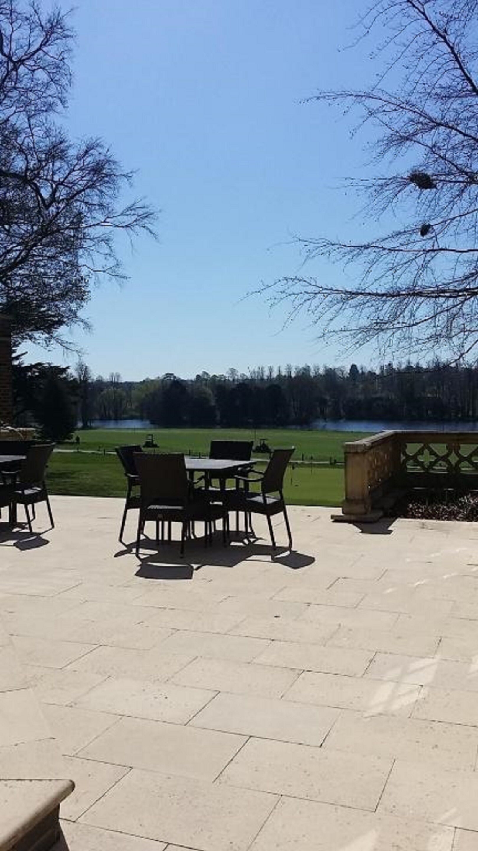 An outdoor patio with a table and chairs, a stone bench, and a view of a lake and trees.