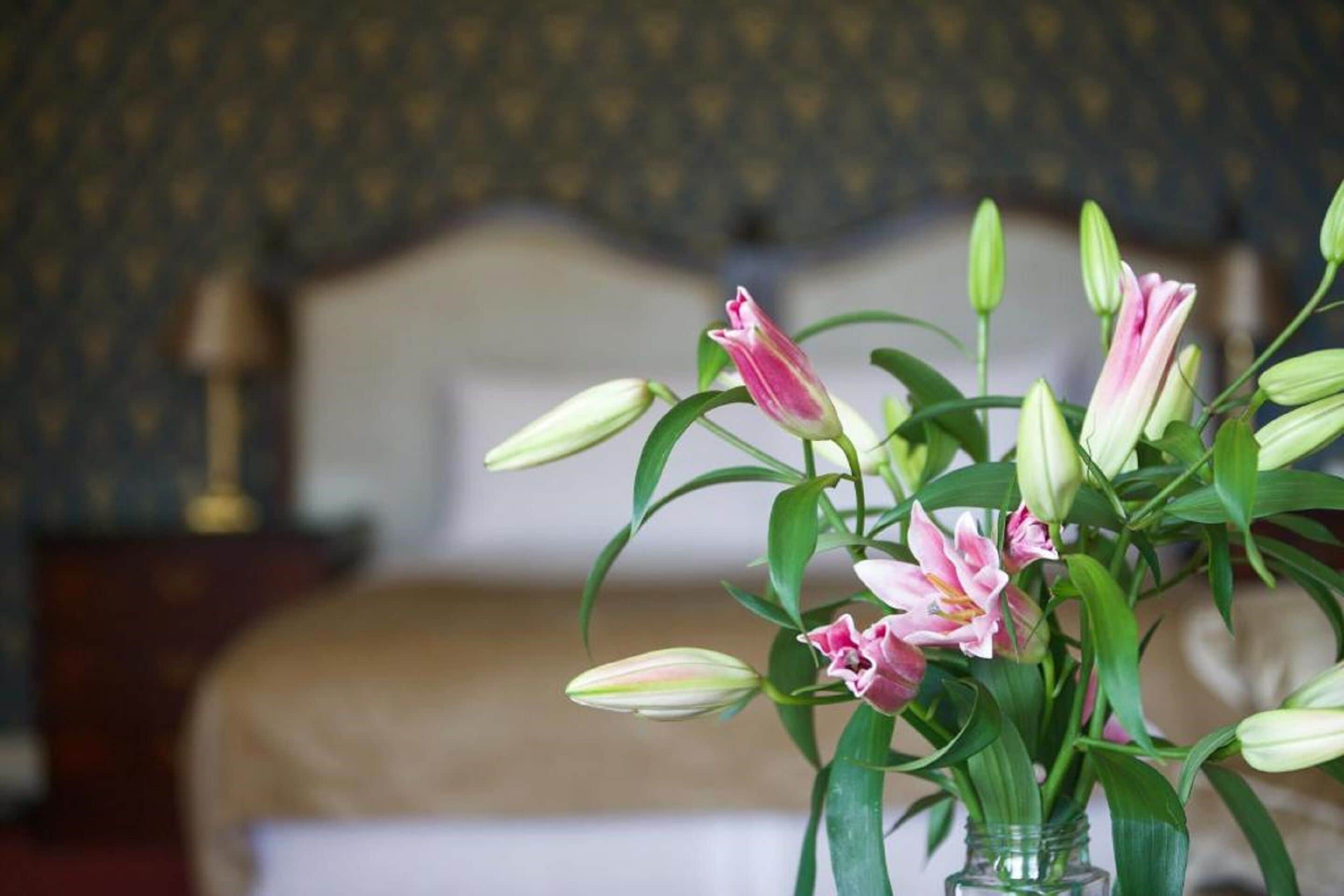 A bouquet of pink and white flowers in a clear glass vase on a bed with a headboard and a lamp in the background.