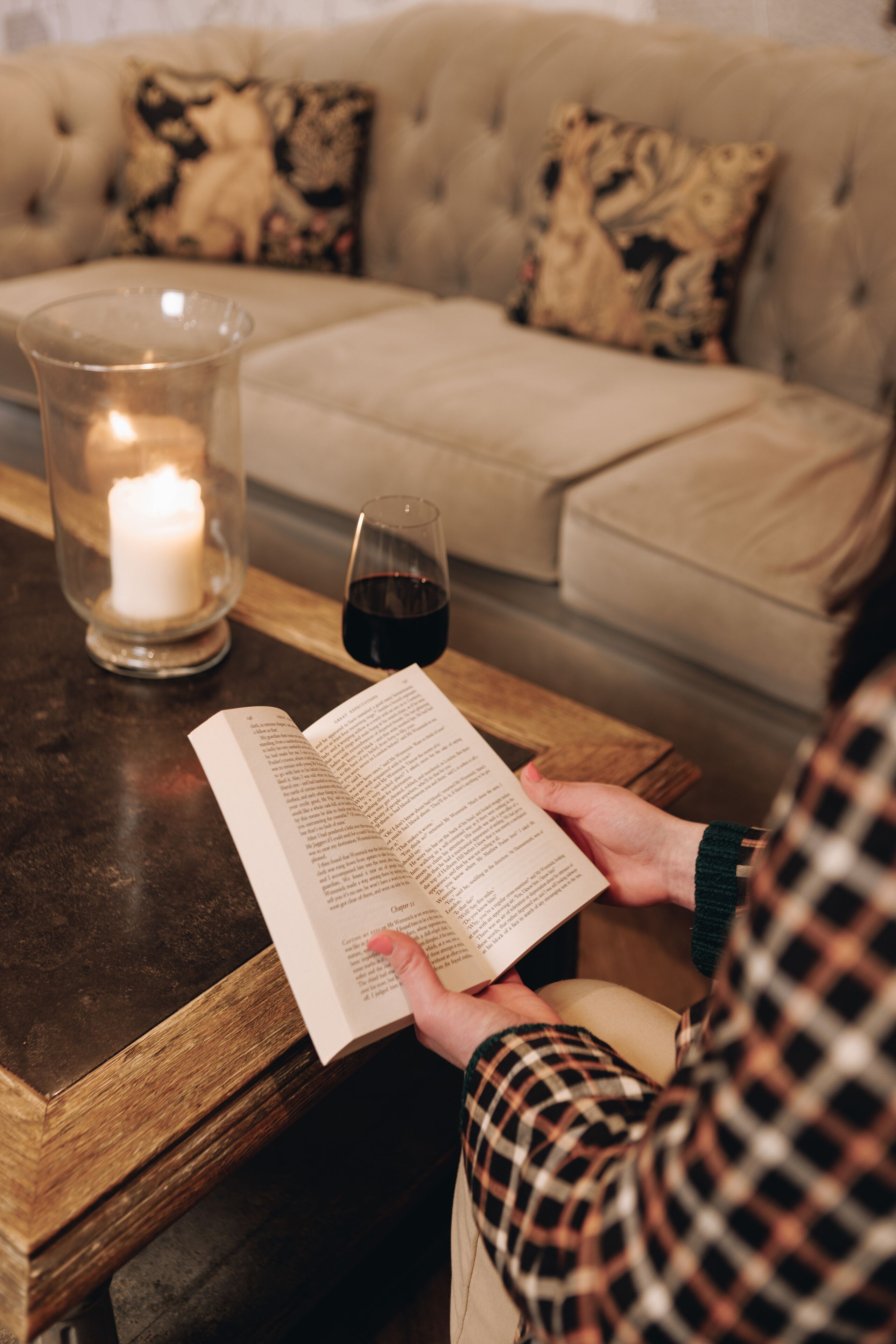 A person reading a book at a wooden table with a candle and a glass of red wine.