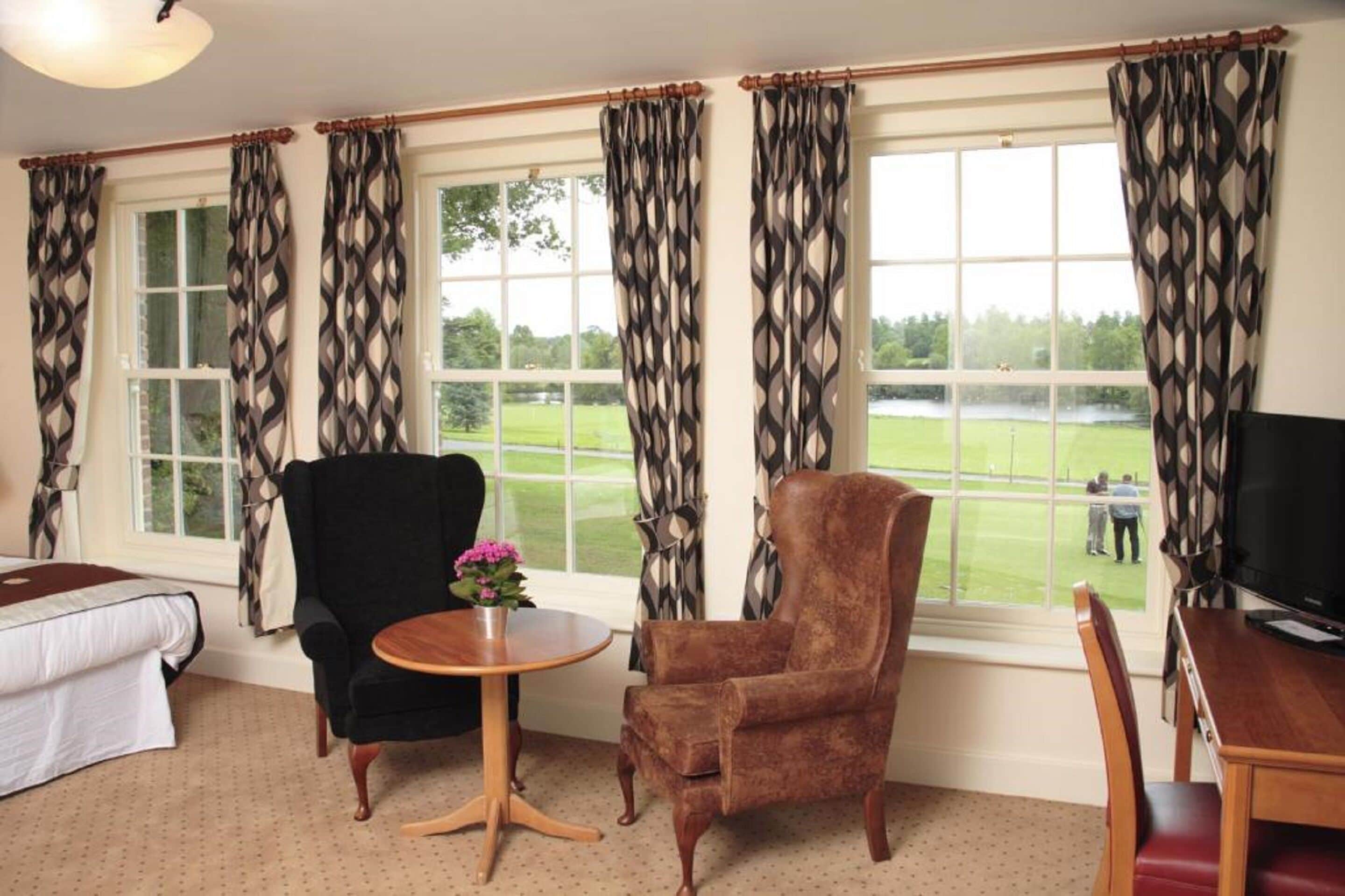 Una habitación con una cama, dos sillones, una pequeña mesa redonda con un florero y un televisor. Amplias ventanas con cortinas estampadas ofrecen vistas a un campo verde.