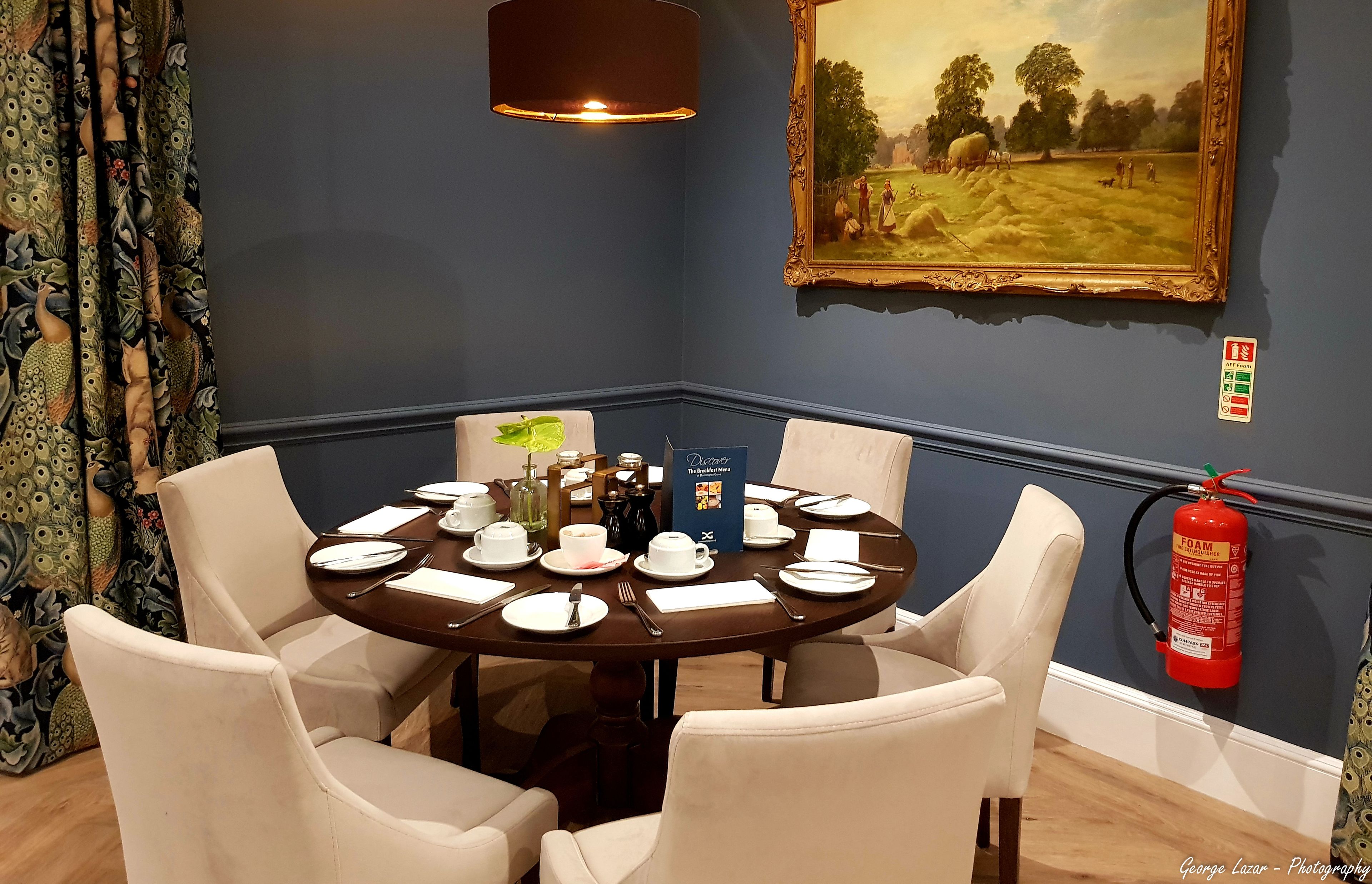 A dining room with a round table set for four, a painting on the wall, and a fire extinguisher mounted on the wall.