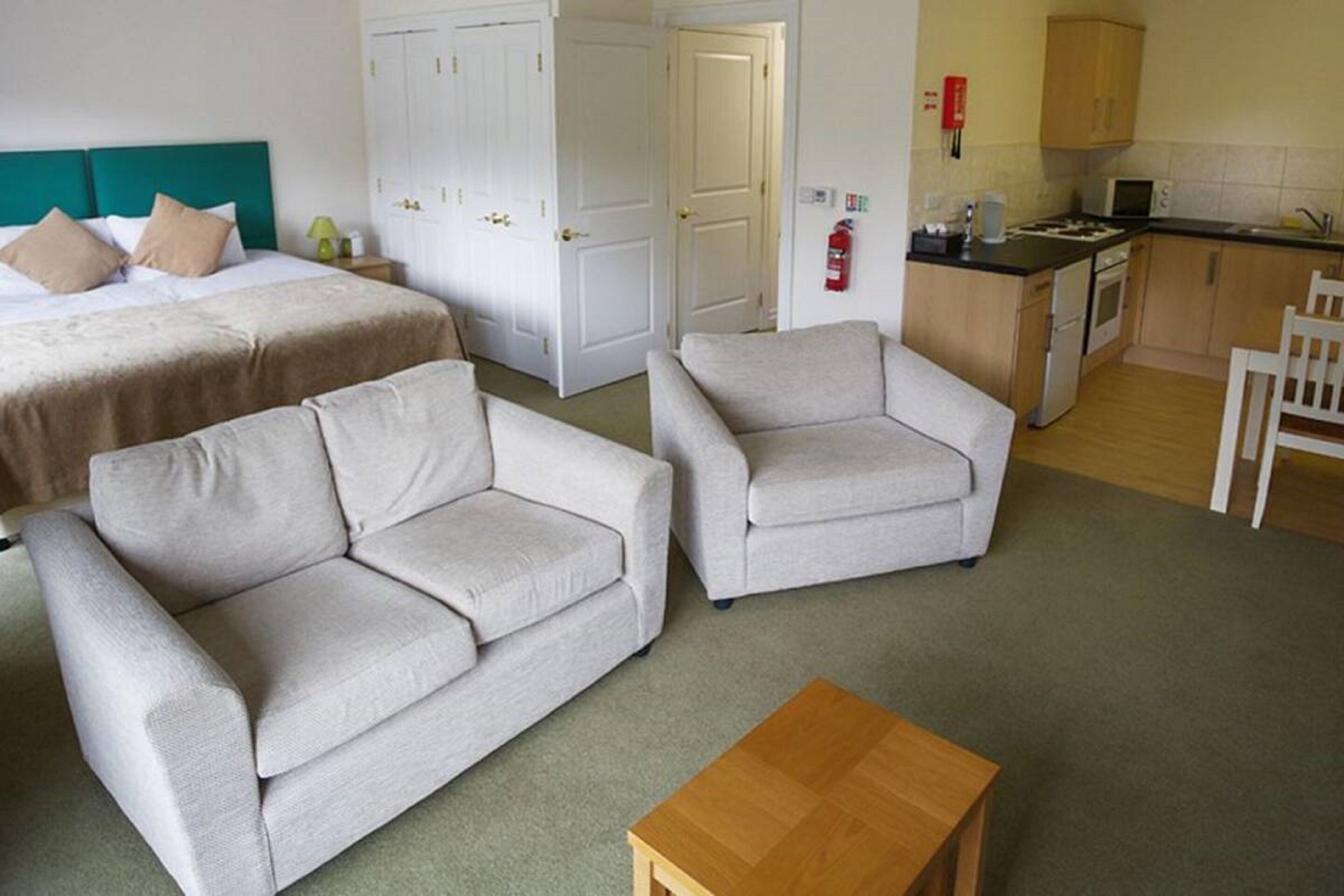 Habitación de hotel con cama, dos sofás, una mesa de centro de madera, kitchenette y una puerta blanca.
