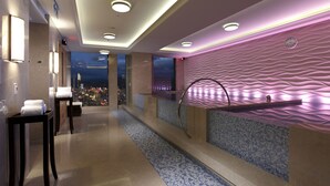 Un moderno espacio de spa con vistas a un paisaje urbano nocturno, una camilla de masaje y un mostrador de recepción con toallas.