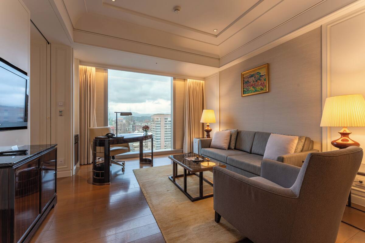 Une chambre d’hôtel moderne équipée d’un canapé, d’un fauteuil, d’une table basse, d’une télévision et offrant une vue sur la ville.