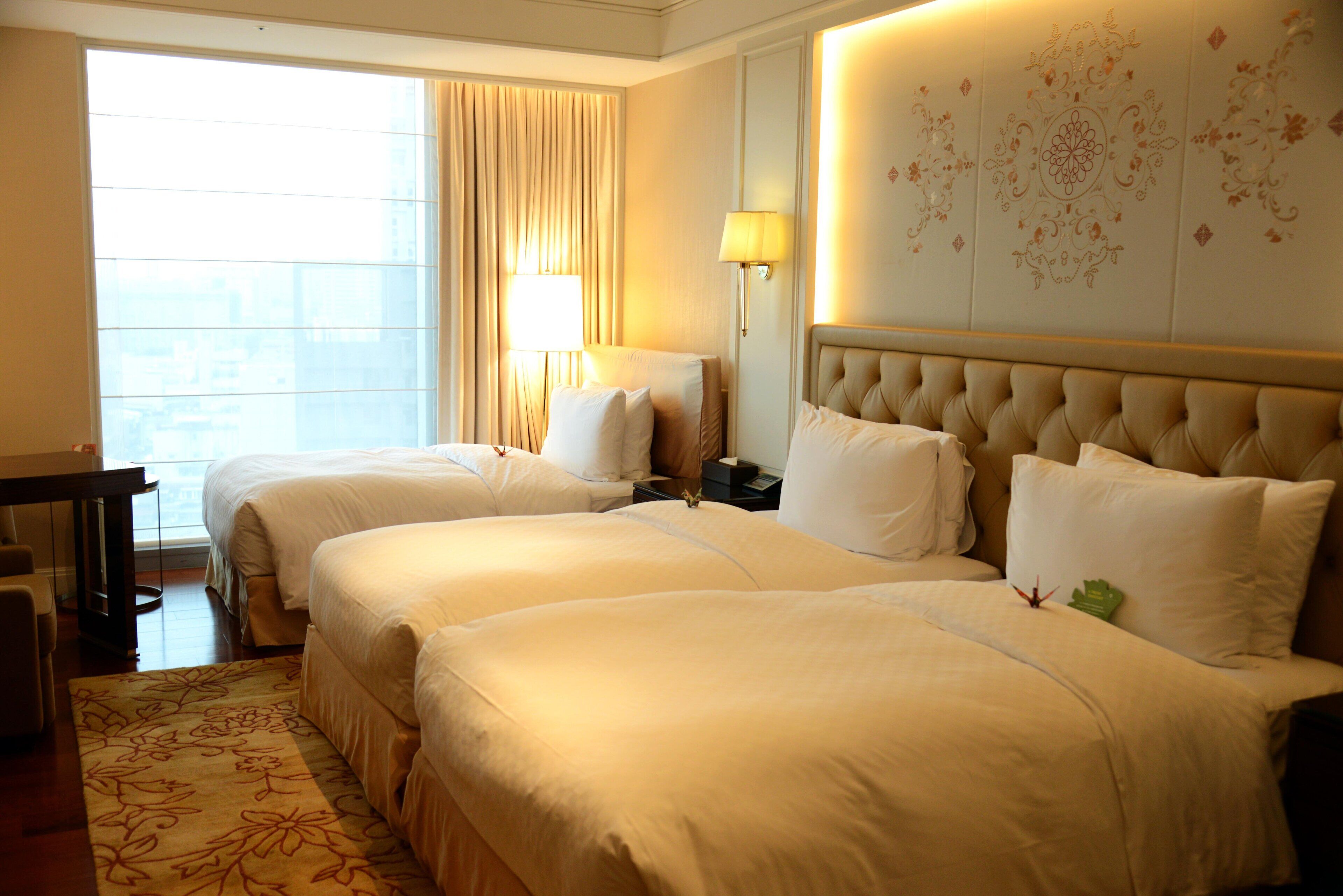 A hotel room with two beds, a desk, a lamp, and a large window with curtains.