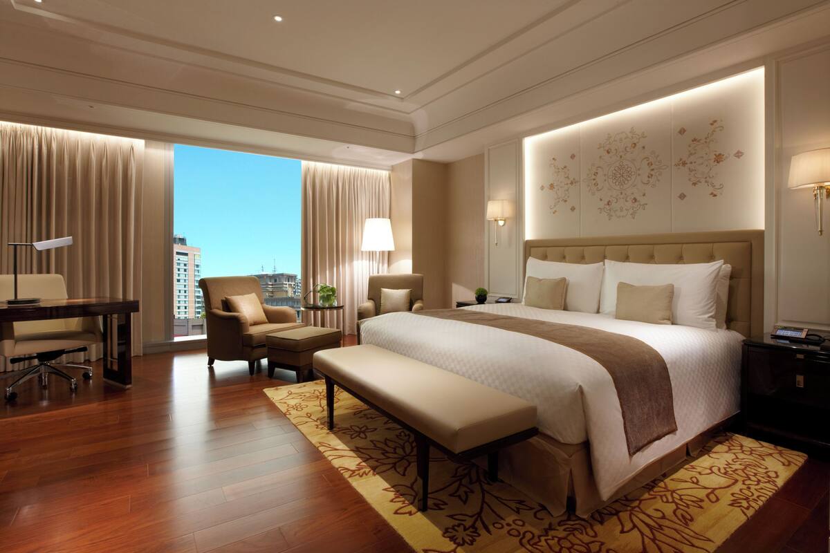 Une chambre d’hôtel avec un grand lit, un bureau, une chaise, un banc et une vue sur la ville.