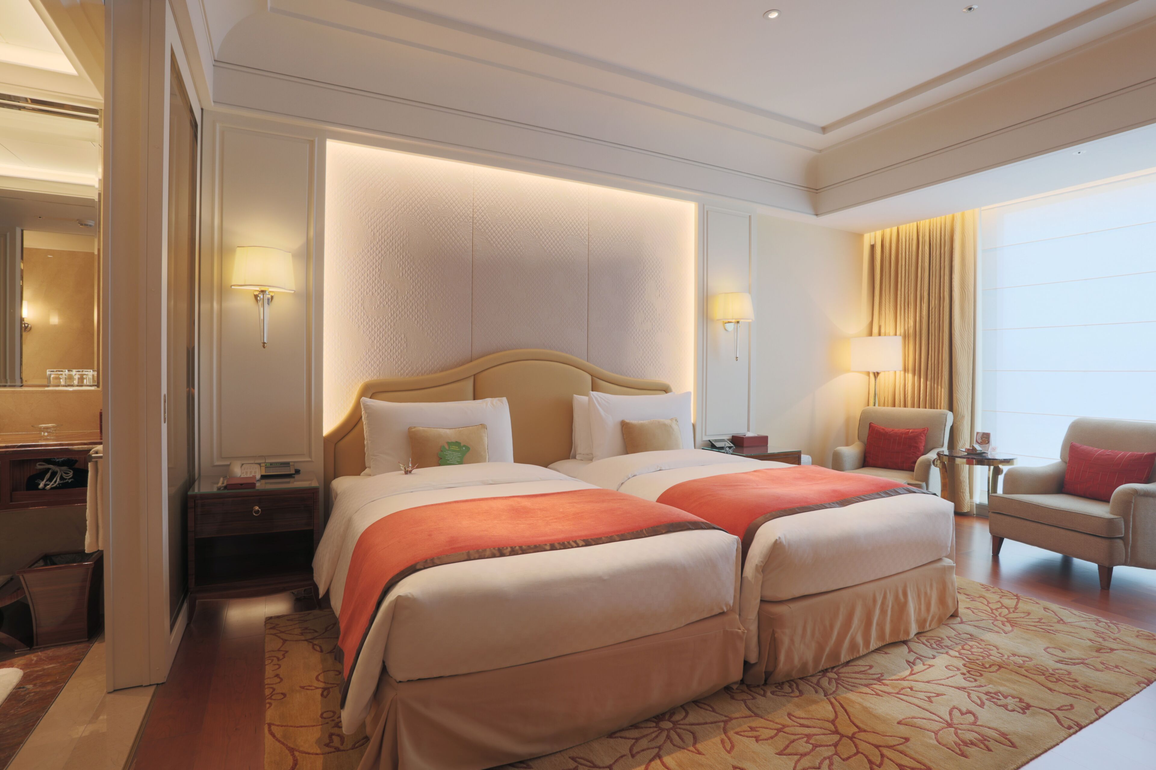 A hotel room with two beds, a sitting area with a chair, a small table, and a large window with curtains.