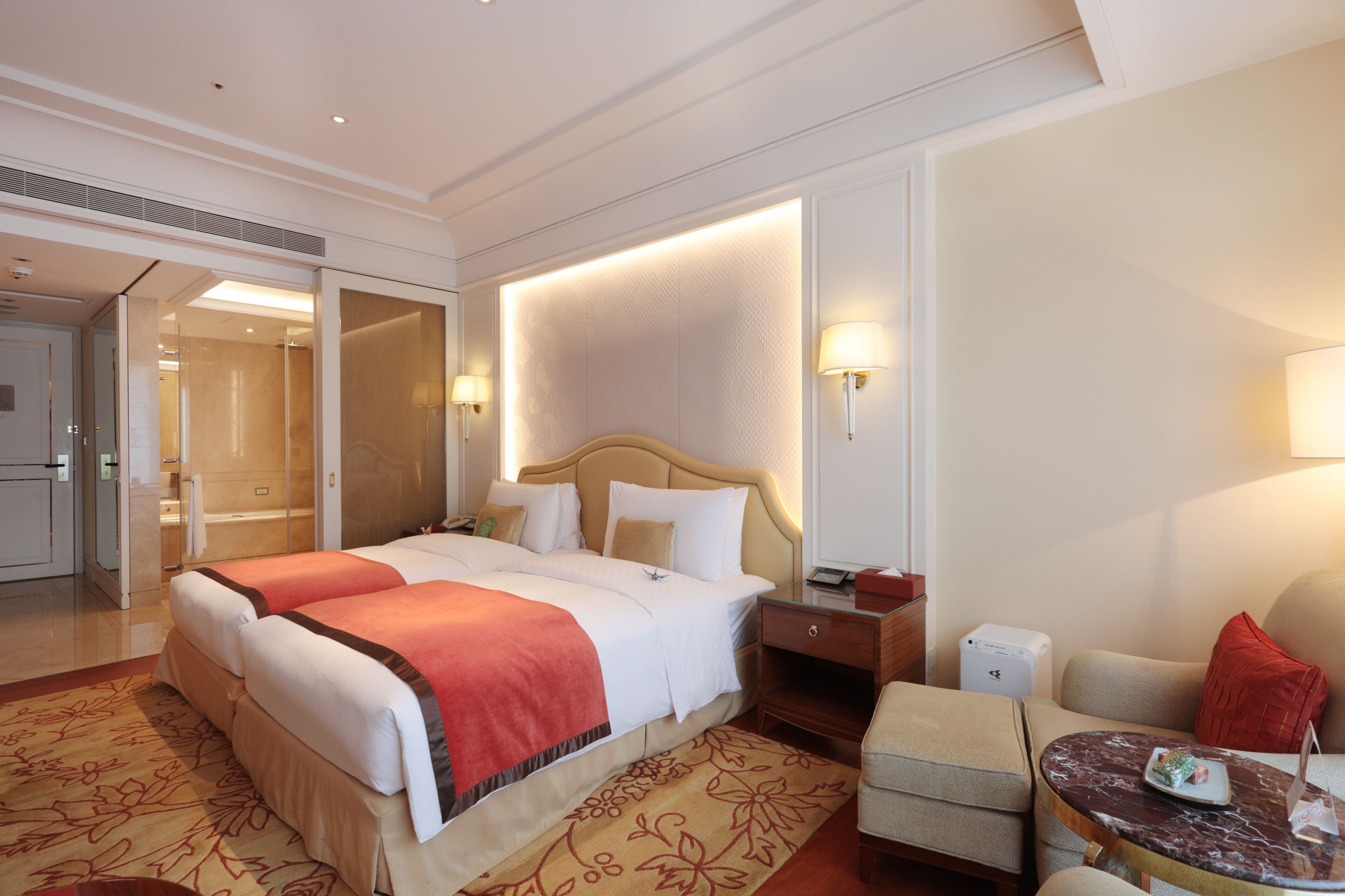 A hotel room with a large bed, bedside tables, a seating area with a chair and a small table, and a bathroom visible through an open door.